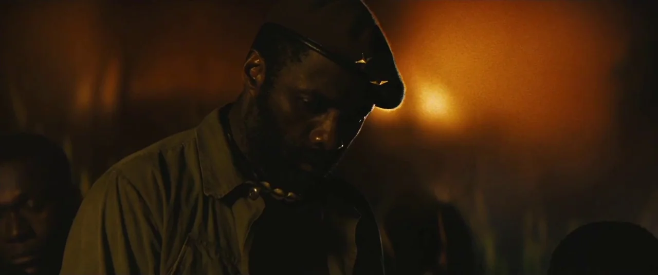 Beasts of No Nation (2015) movie still: medium — A man in a military beret stands at night in a dark outdoor location. Golden light from a fire…