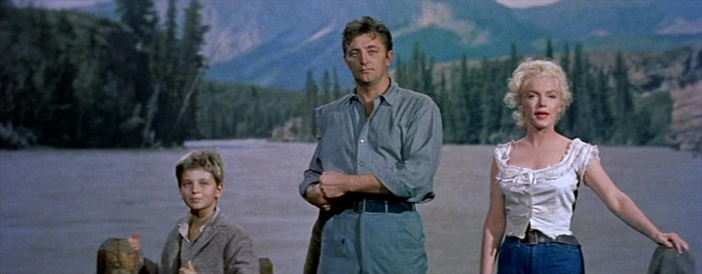 River of No Return (1954) movie still: fullbody — A wide river flows in the background beneath hazy green mountains and pine trees. An adult man, an…