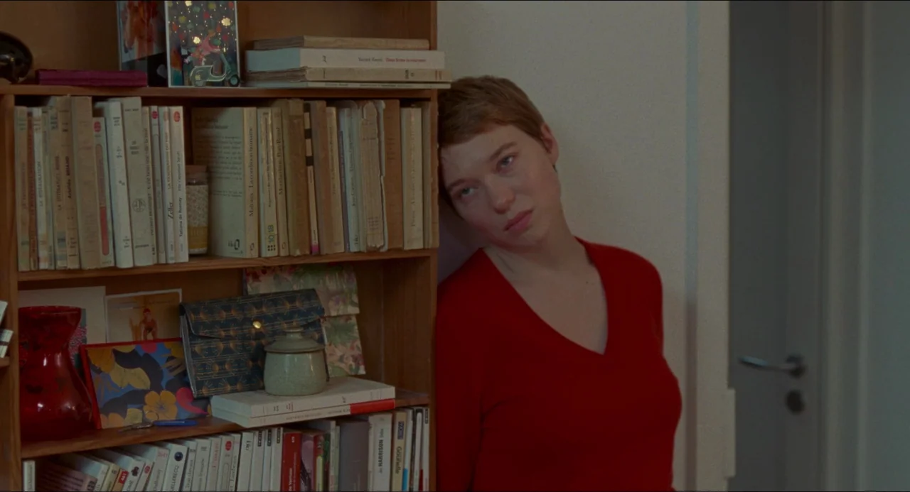 One Fine Morning (2022) movie still: medium — A light-colored wall stands behind a person leaning against a full wooden bookshelf. The person wears…