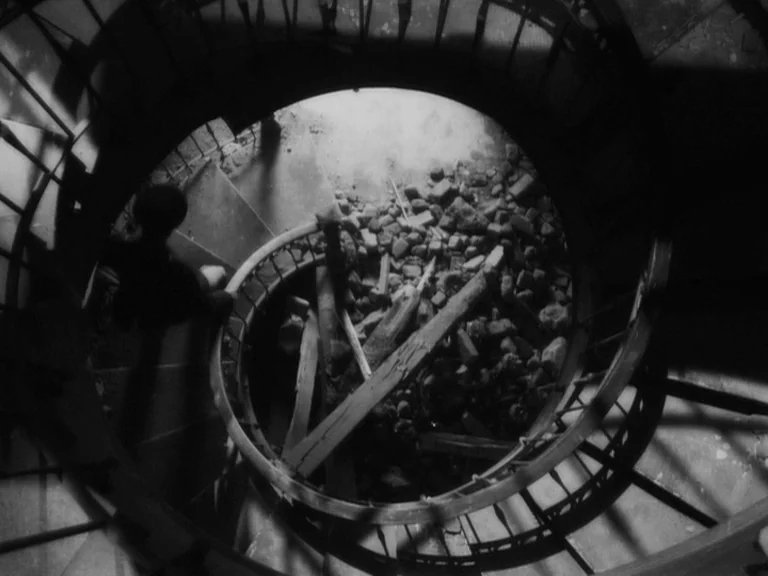 The Good German movie still: wide — A top-down view of a spiral staircase cluttered with wooden beams and stone rubble. The structure is illuminated…