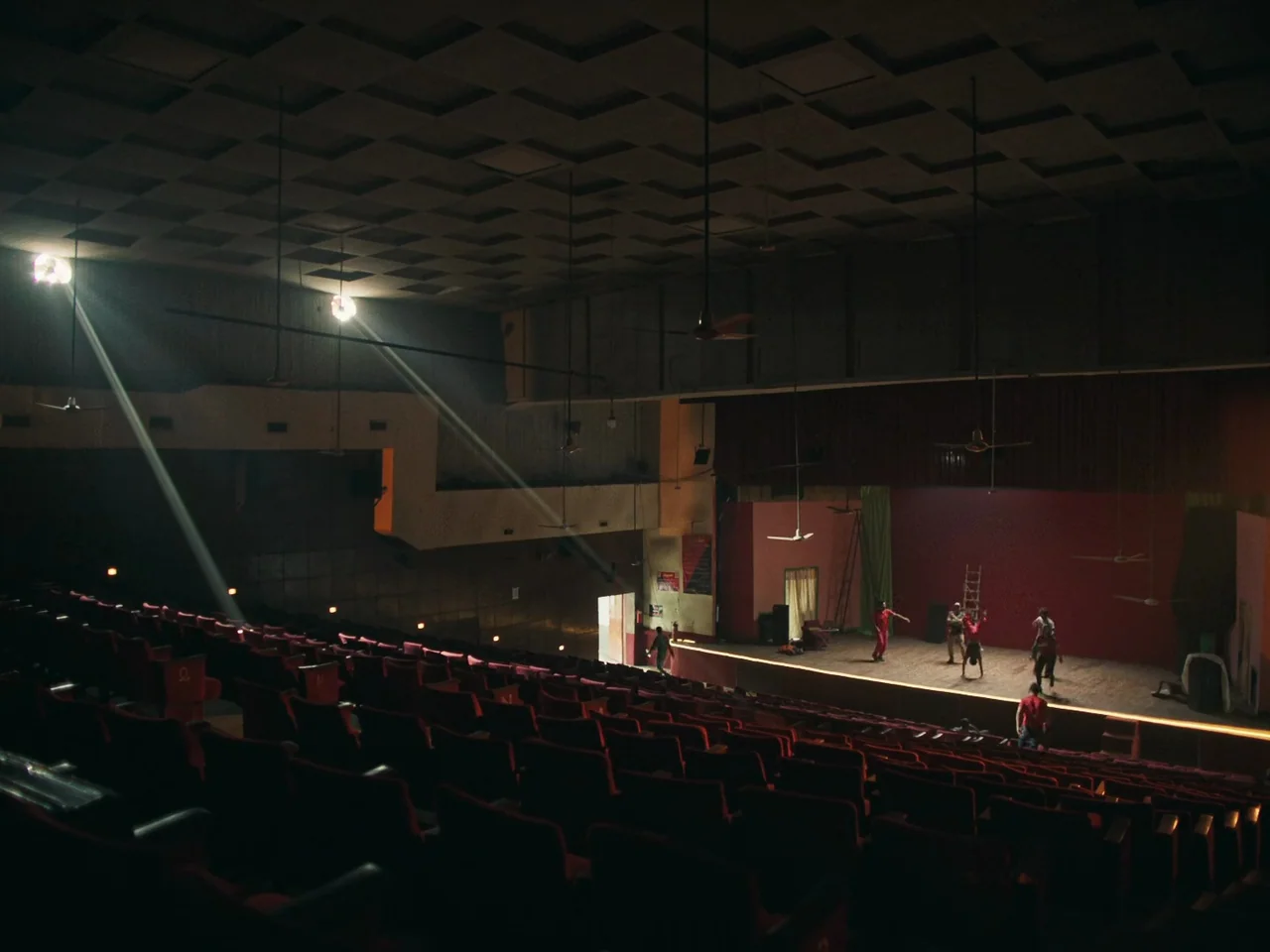 Joyland (2022) movie still: wide — A large dark auditorium features rows of empty red seats under a high ceiling. A group of people practice…