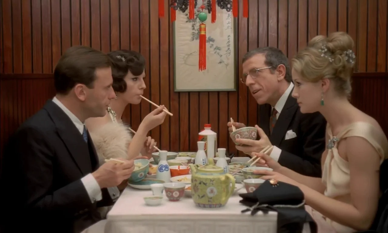 The Conformist (1970) movie still: medium — A group of four people sits at a white-clothed table eating Chinese food. The background features…