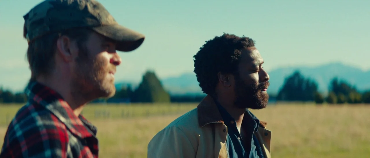 Z for Zachariah (2015) movie still: medium — A vast, open field is set against a backdrop of distant blue-toned mountains. Two men stand in the…