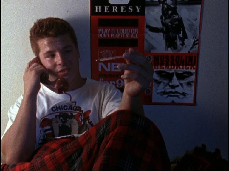 Totally F***ed Up (1993) movie still: medium — A teenager sits in a dim bedroom against a wall adorned with band posters. He holds a phone to his…