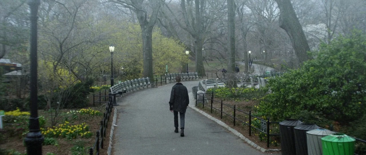 Depraved (2019) movie still: wide — A paved path winds through a park filled with bare trees and blooming yellow flowers. A single person walks…
