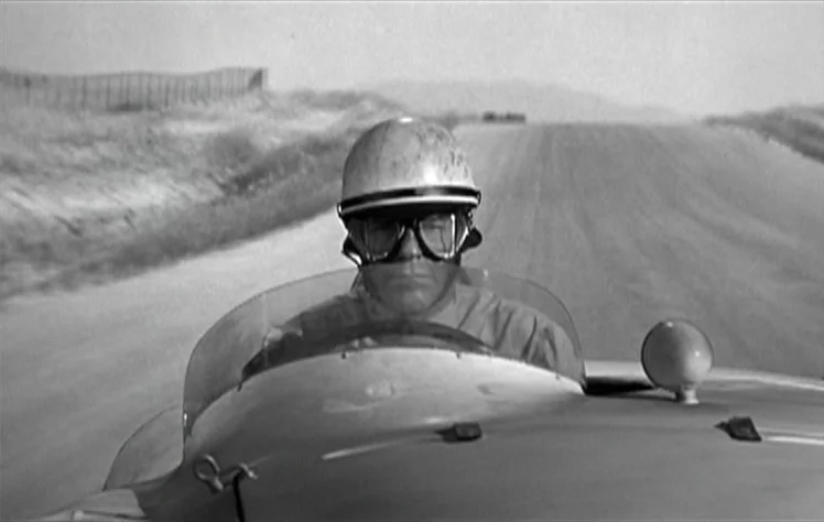 On The Beach (1959) movie still: medium — A vintage race car travels down an open track under bright daylight conditions. The male driver looks…
