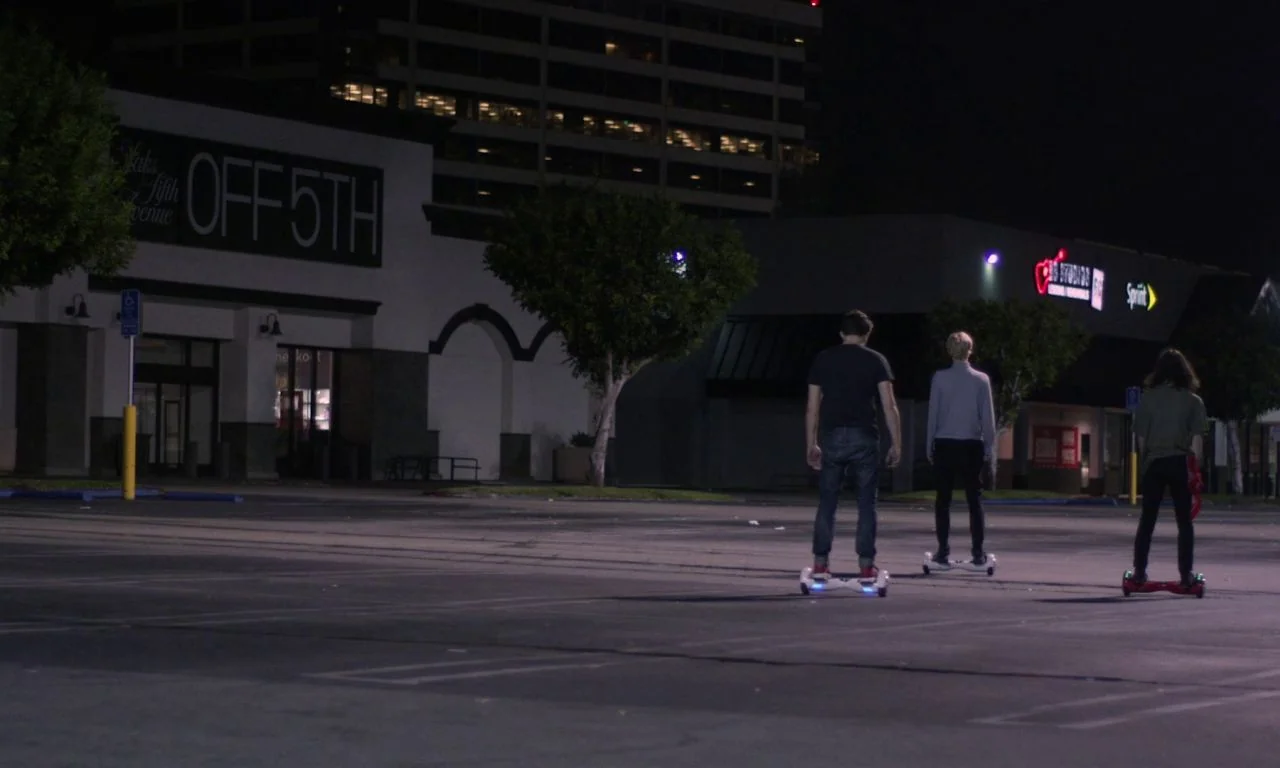 Ham on Rye (2019) movie still: wide — An expansive, empty retail store parking lot at night with illuminated storefront signs visible. Three young…