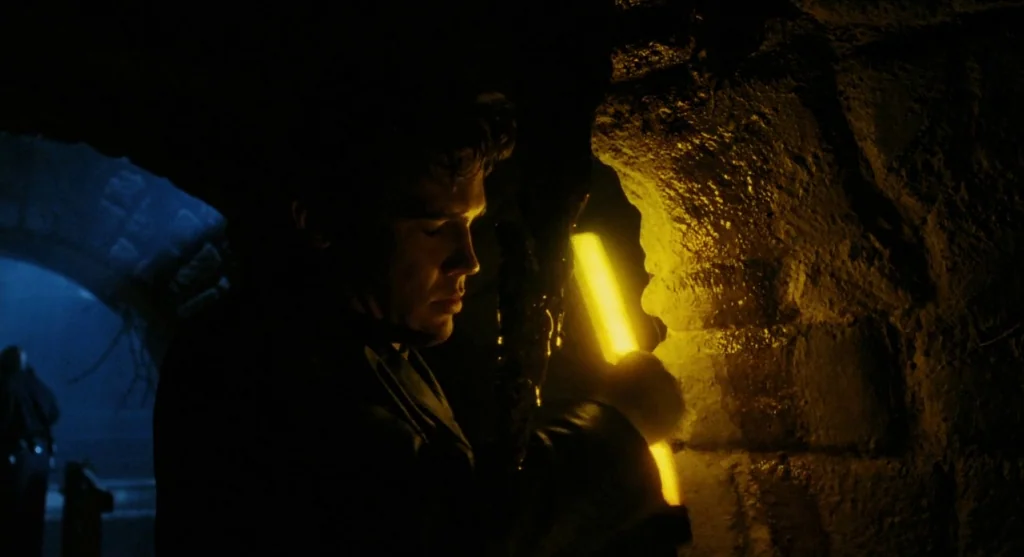 Mimic (1997) movie still: medium — A dark, cramped underground tunnel made of rough, wet stone walls. A man in a leather jacket holds a glowing…