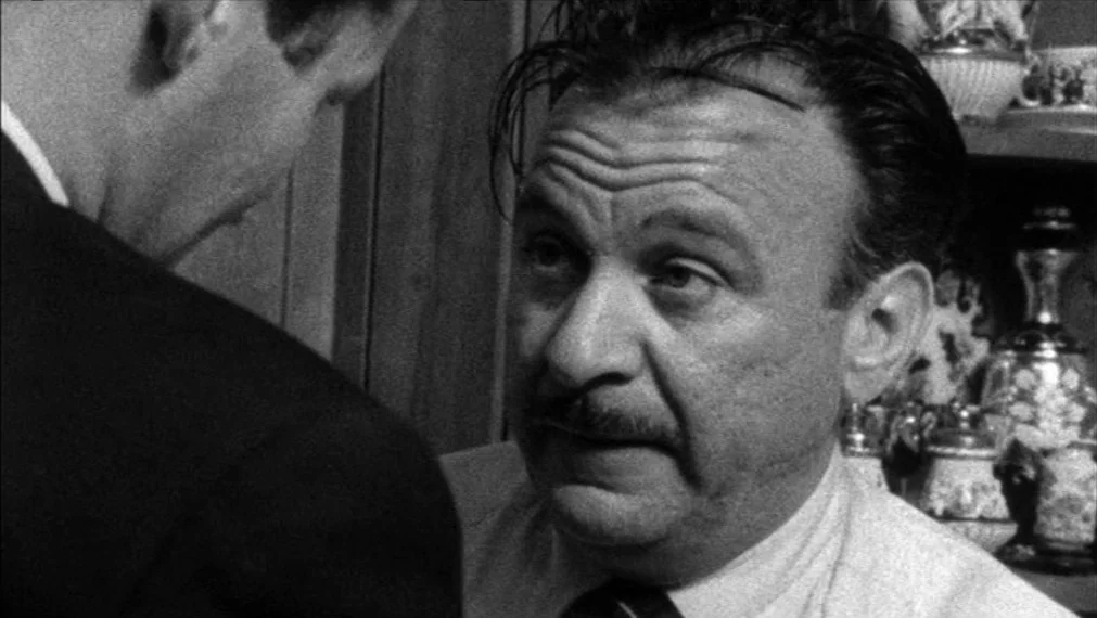 Faces (1968) movie still: closeup — A man in a shirt and tie looks toward an unseen person. Ornate decorative glassware sits on a shelf in the…