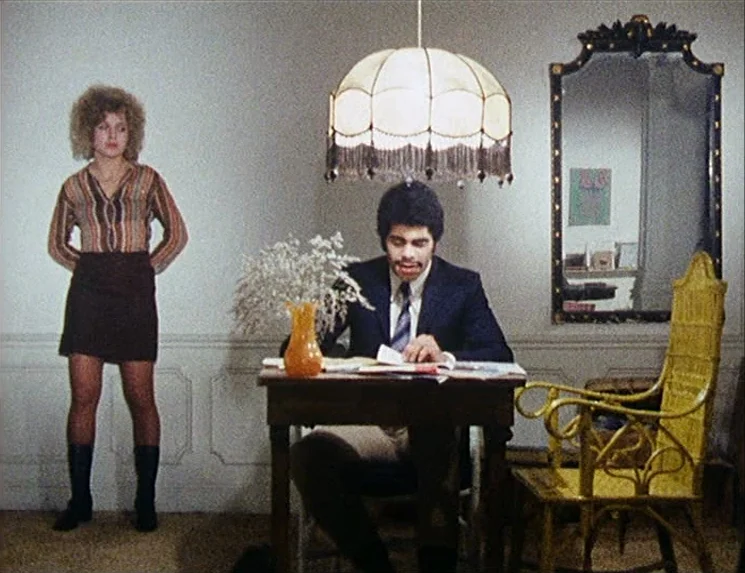 Rio Das Mortes (1971) movie still: fullbody — A sparse room features a plain white wall, a wall mirror, and a yellow chair. A man sits at a table…