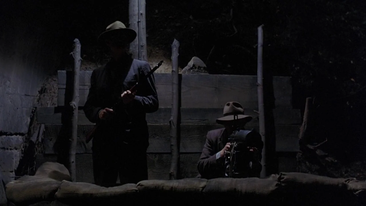 Matewan (1987) movie still: medium — Two men wearing fedoras stand inside a dark, wooden-reinforced military trench at night. They operate weapons…