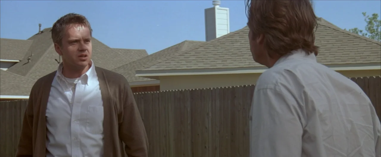 Arlington Road (1999) movie still: medium — A residential backyard with a tall wooden fence and house roofs in the background. Two men face each…
