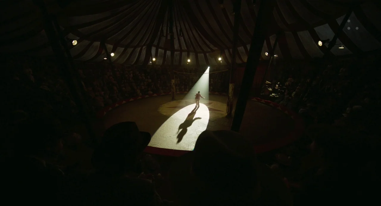Dumbo (2019) movie still: wide — The interior of a darkened canvas circus tent features a central performance ring. A lone performer stands in the…