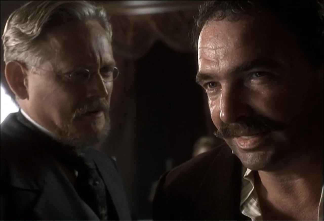 Vendetta (2022) movie still: medium — A dimly lit room with ornate wood paneling visible in the dark background. Two men in period attire stand face…