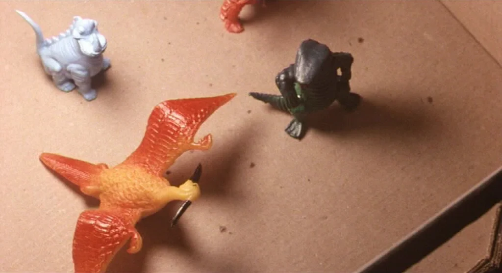King Lear (1987) movie still: wide — Several small plastic dinosaur toys rest on the flat base of a cardboard box. The toys include a blue creature,…