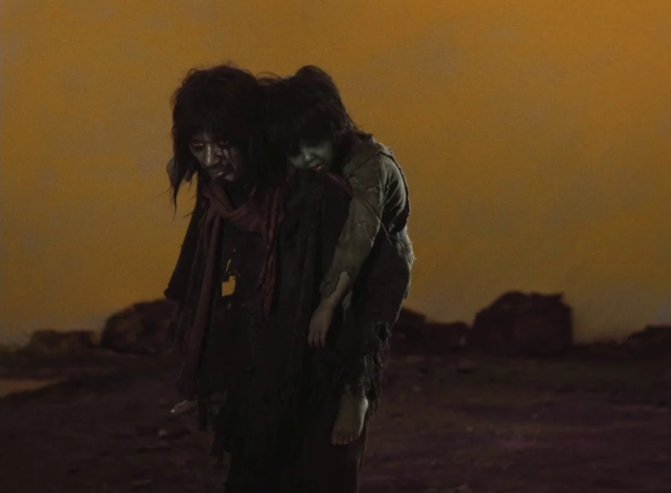 Dodes’ka-den (1970) movie still: fullbody — A desolate, barren landscape stretches beneath an intense, deep-orange, clouded sky. A weary man walks…