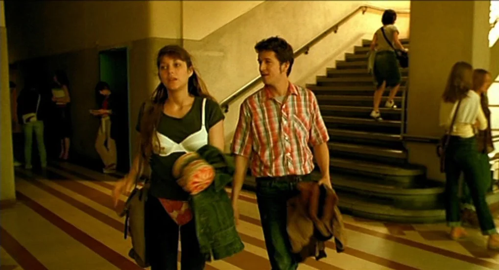 Jeux d’Enfants (2003) movie still: fullbody — A wide stone staircase sits in the background of a large, open school hallway. A young man and woman…