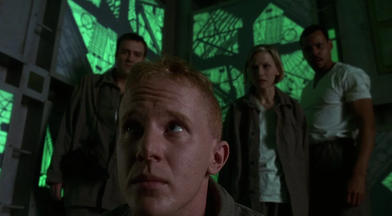 Cube (1997) movie still: medium — Four people stand inside a dark structure made of interconnected green glowing metallic frames. They look upward…