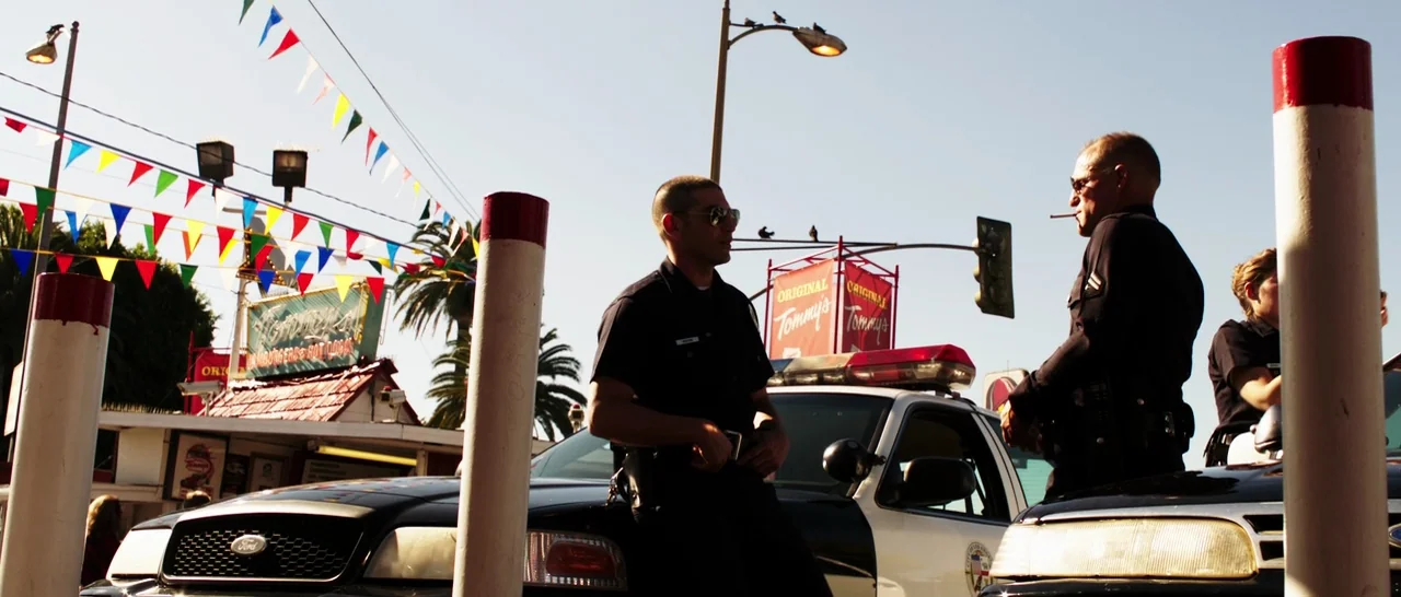 Rampart (2011) movie still: wide — An outdoor parking lot with festive triangular bunting strung between light poles and palm trees. Two uniformed…