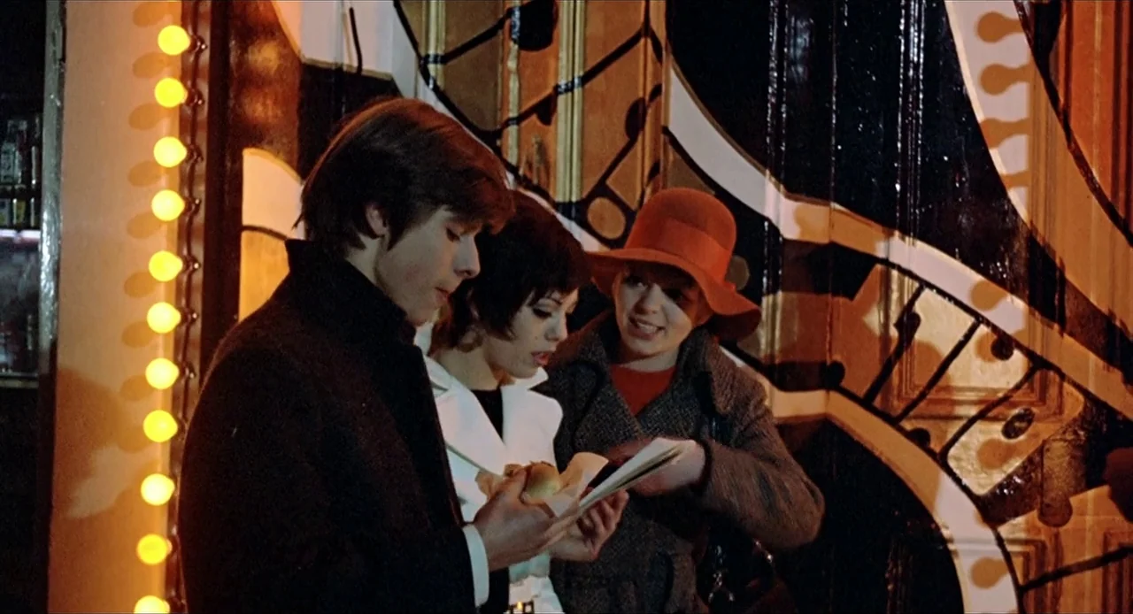 Deep End (1970) movie still: medium — A dimly lit nightclub entrance features a wall with bold black and white graphic patterns. Two young women and…