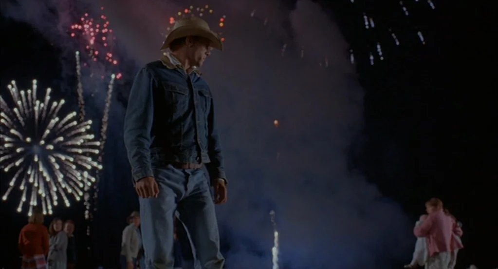 Brokeback Mountain (2005) movie still: fullbody — A nighttime outdoor gathering features large fireworks displays erupting in the dark sky behind…