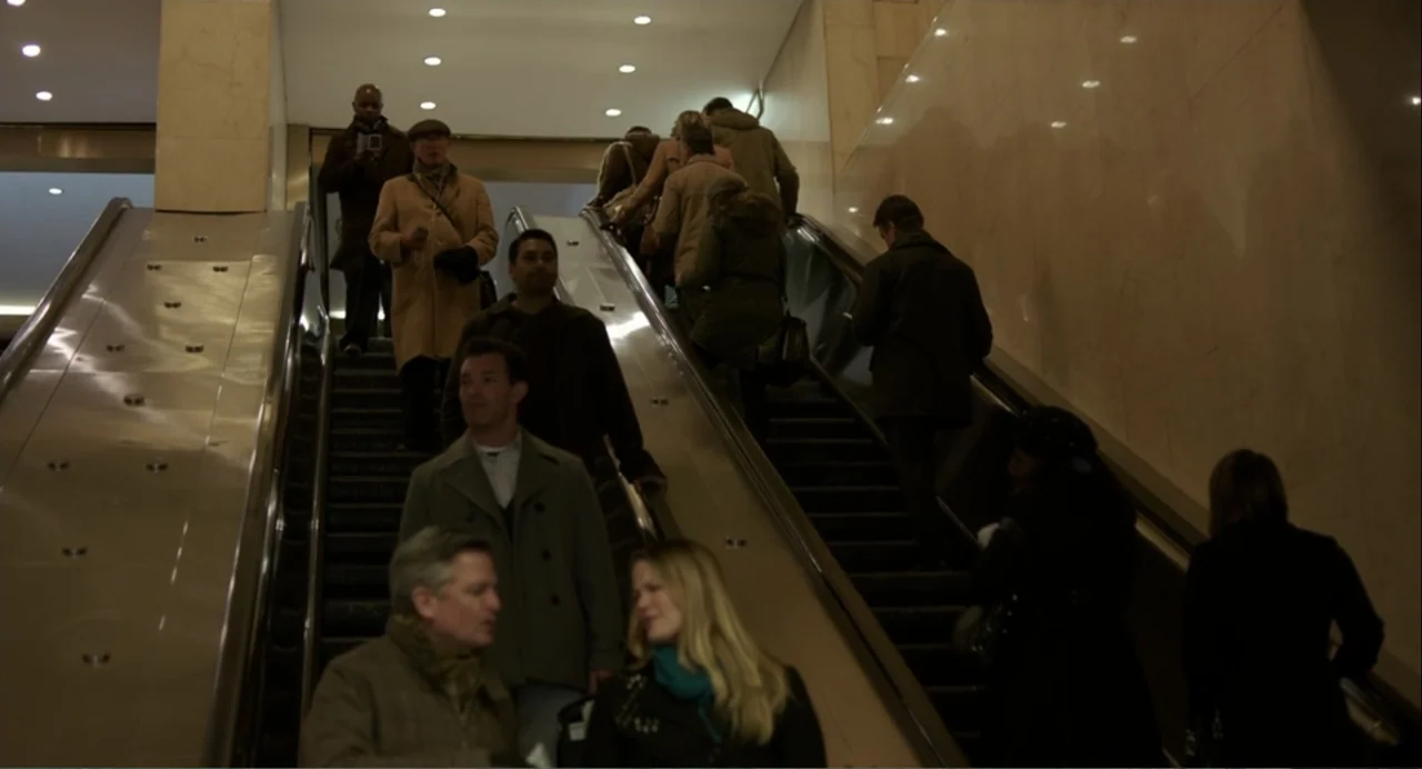 Norman: The Moderate Rise and Tragic Fall of a New York Fixer (2016) movie still: medium — A public indoor escalator moves commuters upward through…