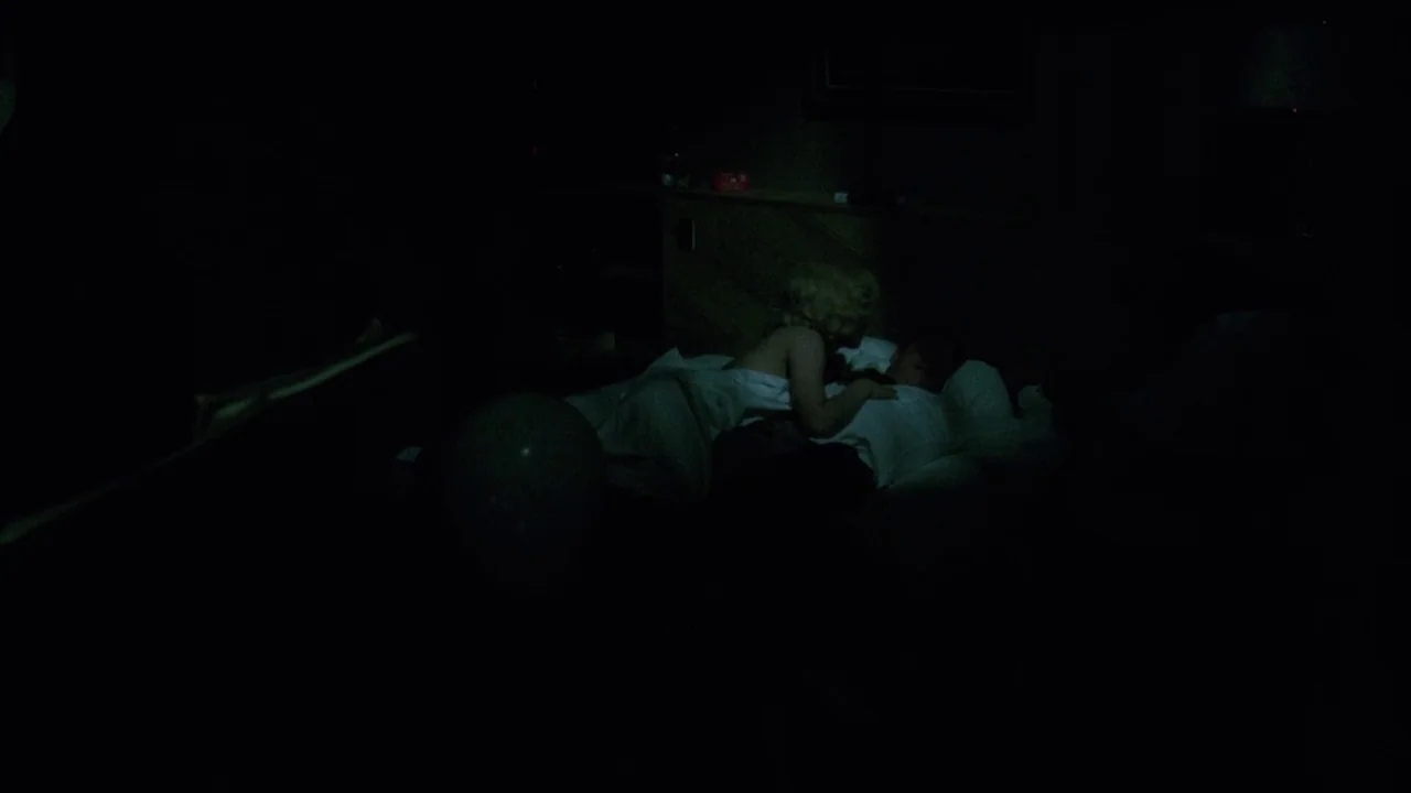 Insignificance (1985) movie still: wide — A dark hotel room with a couple lying together in a bed. The figures are partially illuminated against the…