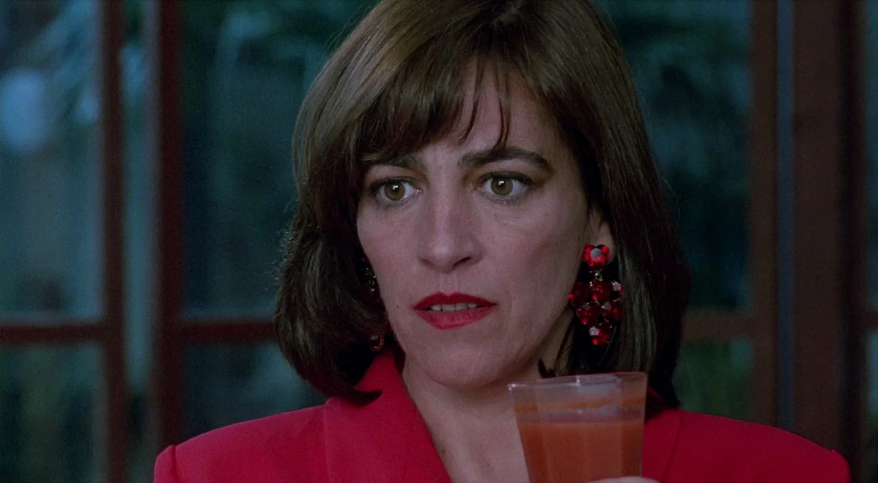 Women on the Verge of a Nervous Breakdown (1988) movie still: closeup — A woman in a red blazer stands in a dim restaurant with large windows behind…
