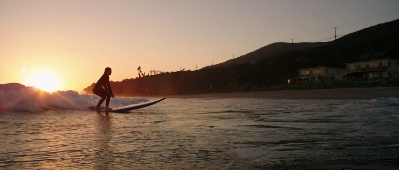 Wish I Was Here (2014) movie still: wide — A coastline at sunset with rolling waves and hills in the background. A lone surfer rides a wave in the…