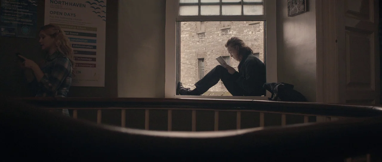 Byzantium (2012) movie still: wide — A stairwell landing features a large window, a wooden handrail, and wall-mounted educational flyers. A man sits…