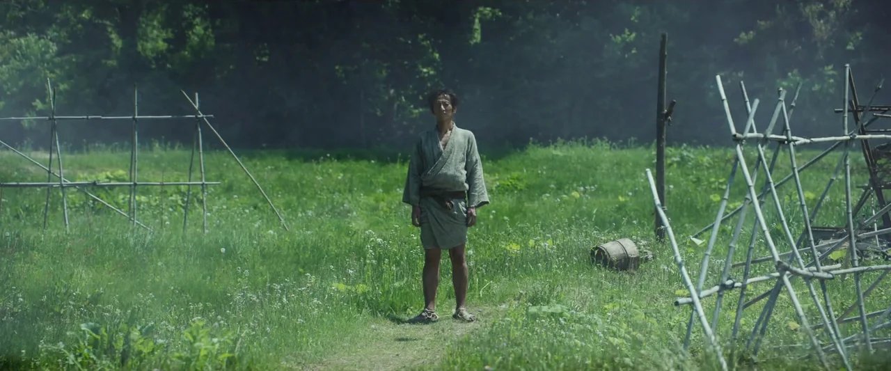 Kubi (2023) movie still: fullbody — A man stands in an open field of tall green grass and wildflowers. Rough bamboo frames and a wooden bucket are…