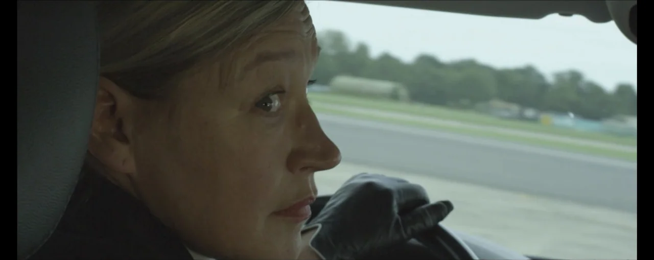 Turbulence (2025) movie still: closeup — A woman drives a car along a road near an airfield. She holds the steering wheel while looking out the side…