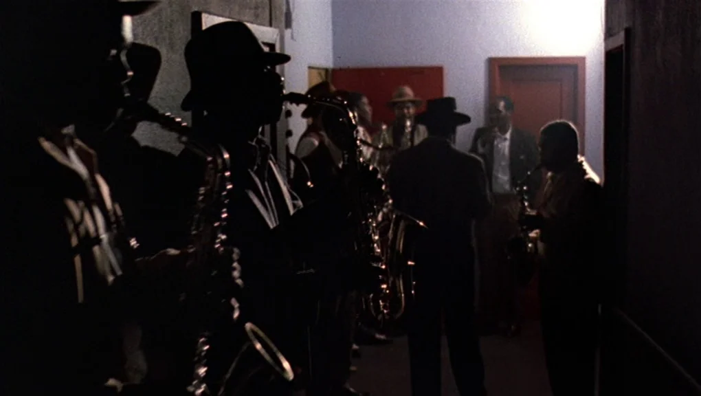 Bird (1988) movie still: medium — A dark, narrow interior hallway features a group of musicians standing in line. The men play saxophones while…