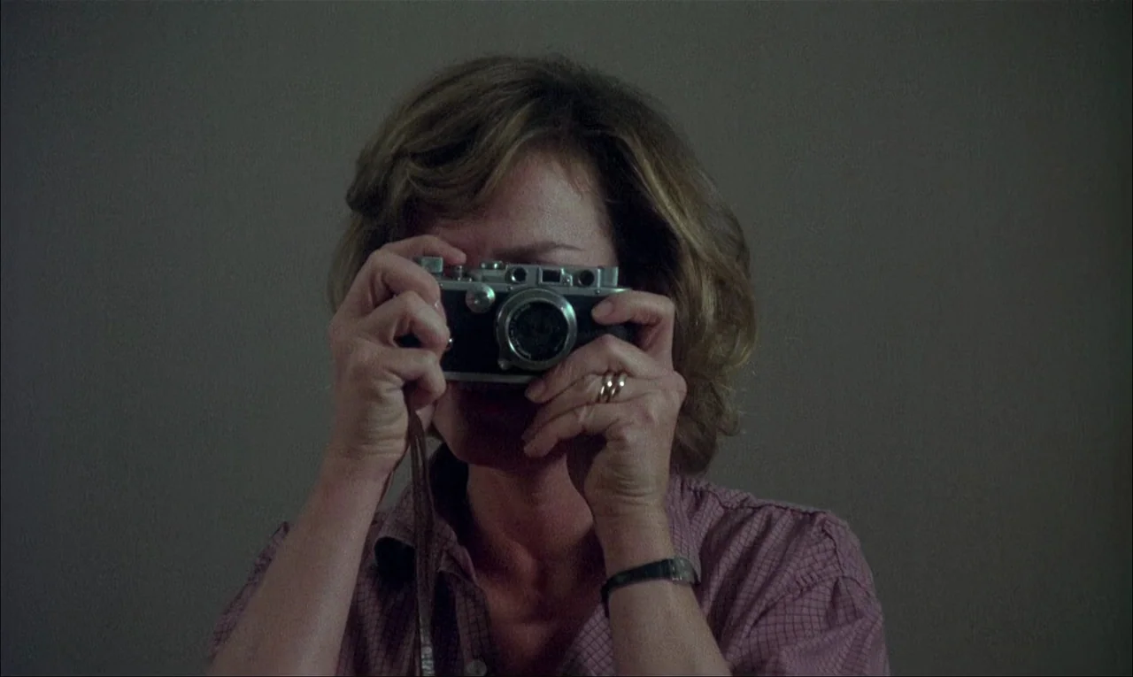 My Life As A Dog (1985) movie still: closeup — A woman stands in a nondescript room with plain, muted grey walls. She holds a vintage 35mm camera to…