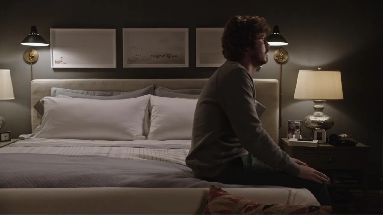 The Intern (2015) movie still: medium — A modern bedroom features a large upholstered headboard and matching bedside tables with lamps. A man sits…