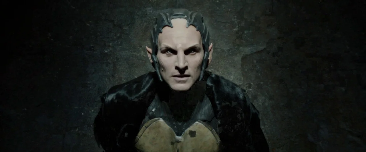 Thor: The Dark World (2013) movie still: closeup — A dark, textured stone wall forms the backdrop for an elf-like male character. The man with pale…