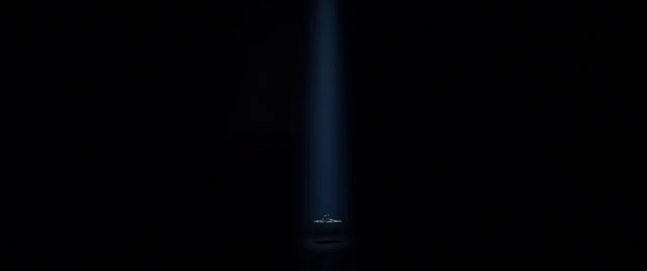 The Platform (2019) movie still: establishing — A dark void contains only a single desk illuminated by a thin vertical spotlight. The desk is…