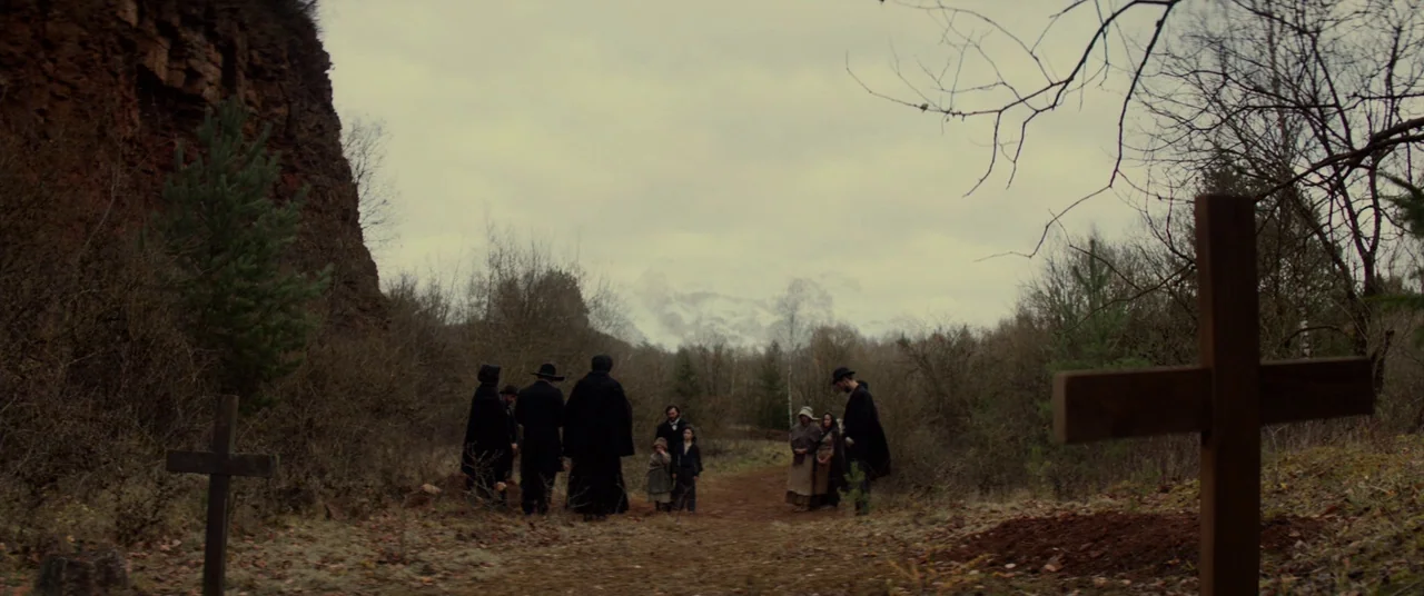 Never Grow Old (2019) movie still: wide — A rural, overgrown cemetery clearing is surrounded by dark bushes and a steep cliff. A small group of…