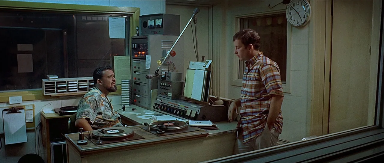 American Graffiti (1973) movie still: medium — A 1970s radio station booth features analog equipment, turntables, and a mounted analog wall clock.…