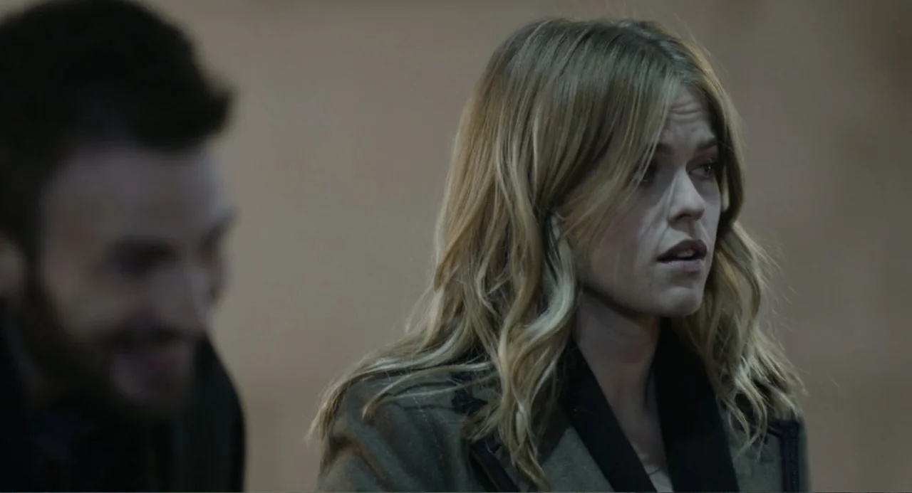 Before We Go (2014) movie still: medium — A dimly lit indoor space with a plain, neutral-colored wall behind the figures. A woman in an olive coat…