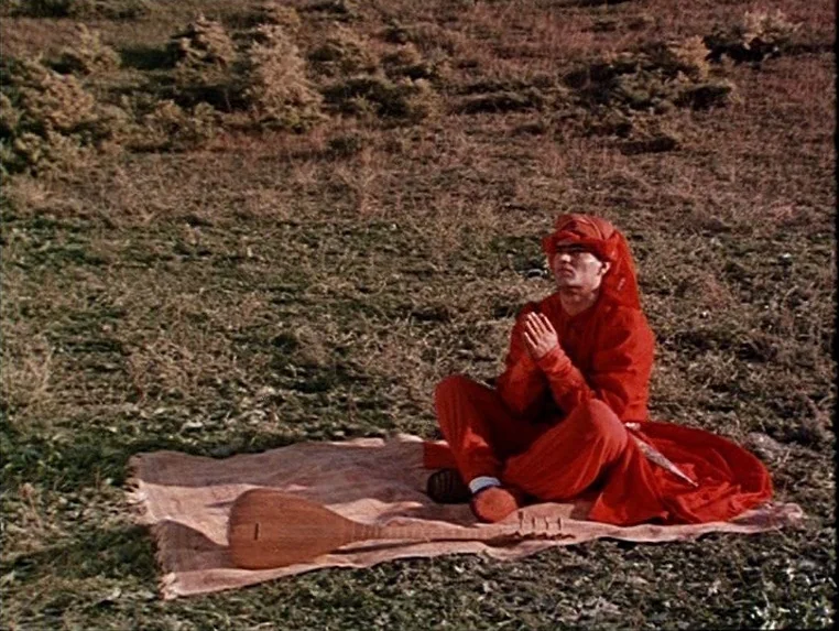 Ashik Kerib (1988) movie still: wide — A man in vivid red attire sits on a patterned cloth in a grassy field. A wooden stringed instrument lies on…