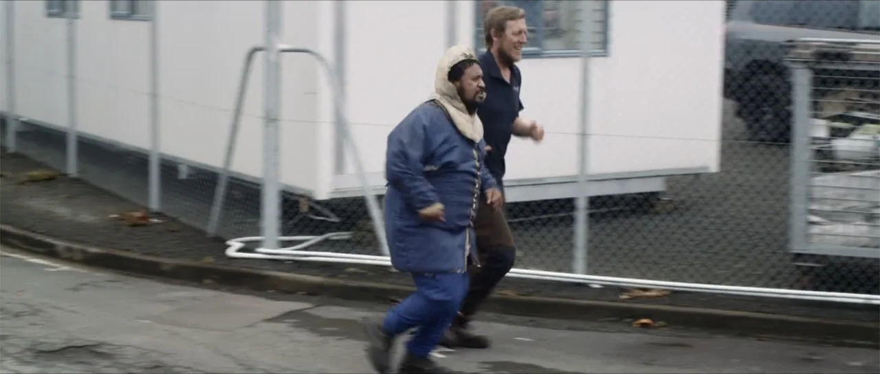 Bludgeon (2018) movie still: fullbody — Two men run along a chain-link fence beside white modular trailers in daylight. One man wears a quilted…
