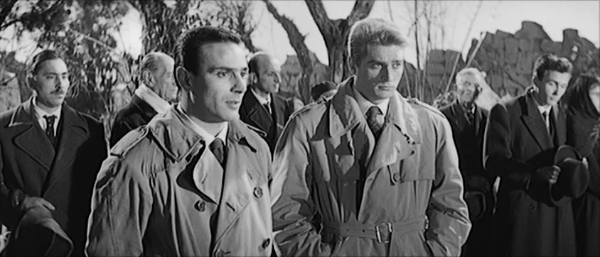 I Vampiri (1957) movie still: medium — A group of men stand together outdoors among bare trees and stone markers. The men wear formal trench coats…