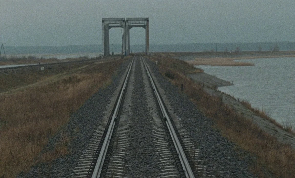 High Life (2018) movie still: establishing — A desolate railway track cuts through a flat landscape adjacent to a large body. Two concrete bridge…