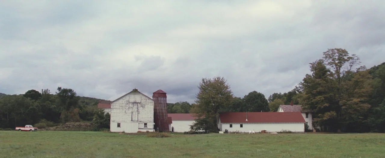 Martha Marcy May Marlene (2011) movie still: wide — A rural landscape features a weathered white barn with a silo next to buildings. A red and white…