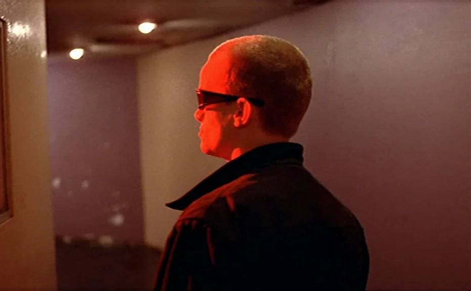 Diva (1981) movie still: medium — A dark hallway with smooth walls and recessed ceiling lights in the background. A man stands in profile wearing…