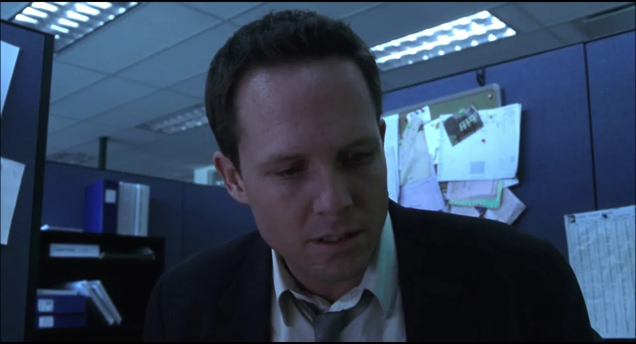 Hellraiser: Hellseeker (2002) movie still: closeup — A man in a suit sits inside a corporate cubicle with navy blue partitions. The man gazes…