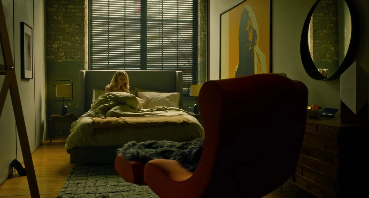 Rabid (2019) movie still: wide — A stylish bedroom features a large window with blinds and an orange pop art portrait. A blonde woman sits alone in…