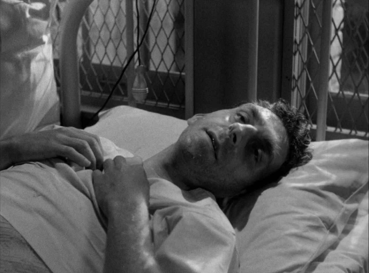 The Killers (1946) movie still: closeup — A hospital bed is positioned against a wall with a window featuring metal grating. A man lies on the bed…