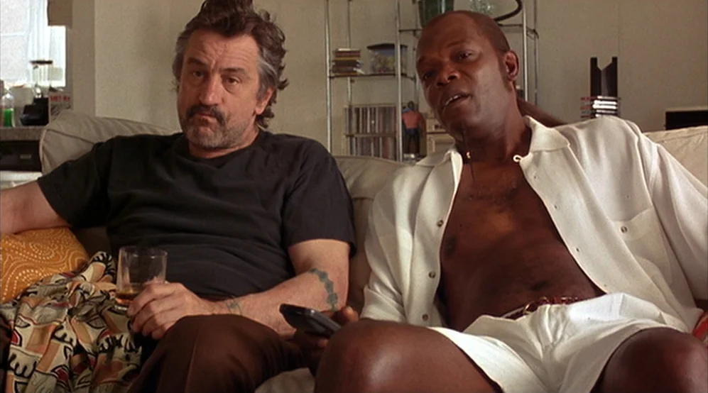 Jackie Brown movie still: medium — A sunny apartment interior features two men relaxing on a beige sofa. The man on the left holds a glass while the…